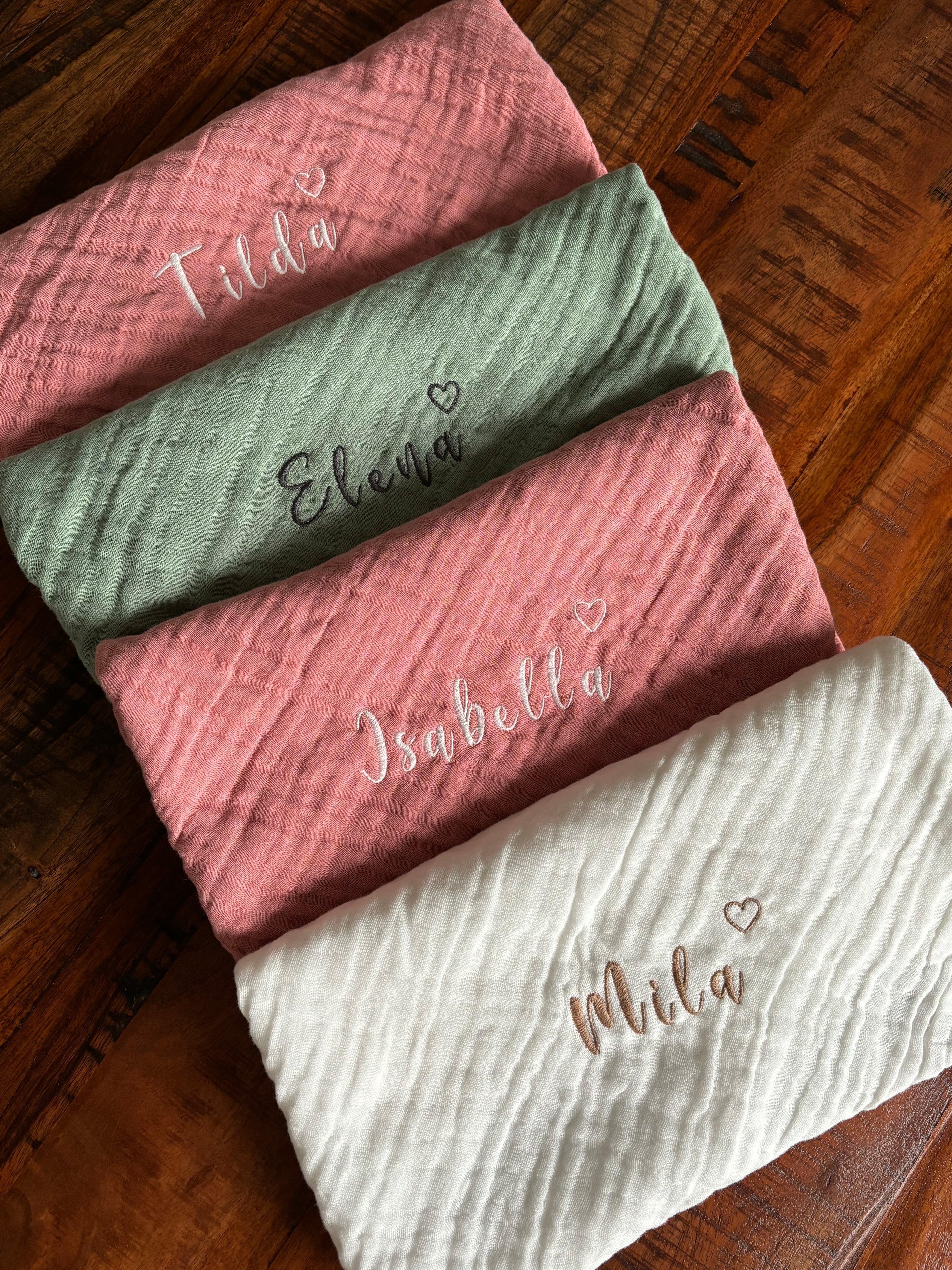 Es sind verschiedene Farben der Musselinstoff Spucktücher abgebildet. Auf jedem Tuch ist eine Namensstickerei, welche individuell und personalisiert abgestimmt ist. Zusätzlich zum Namen ist ein Herz gestickt.