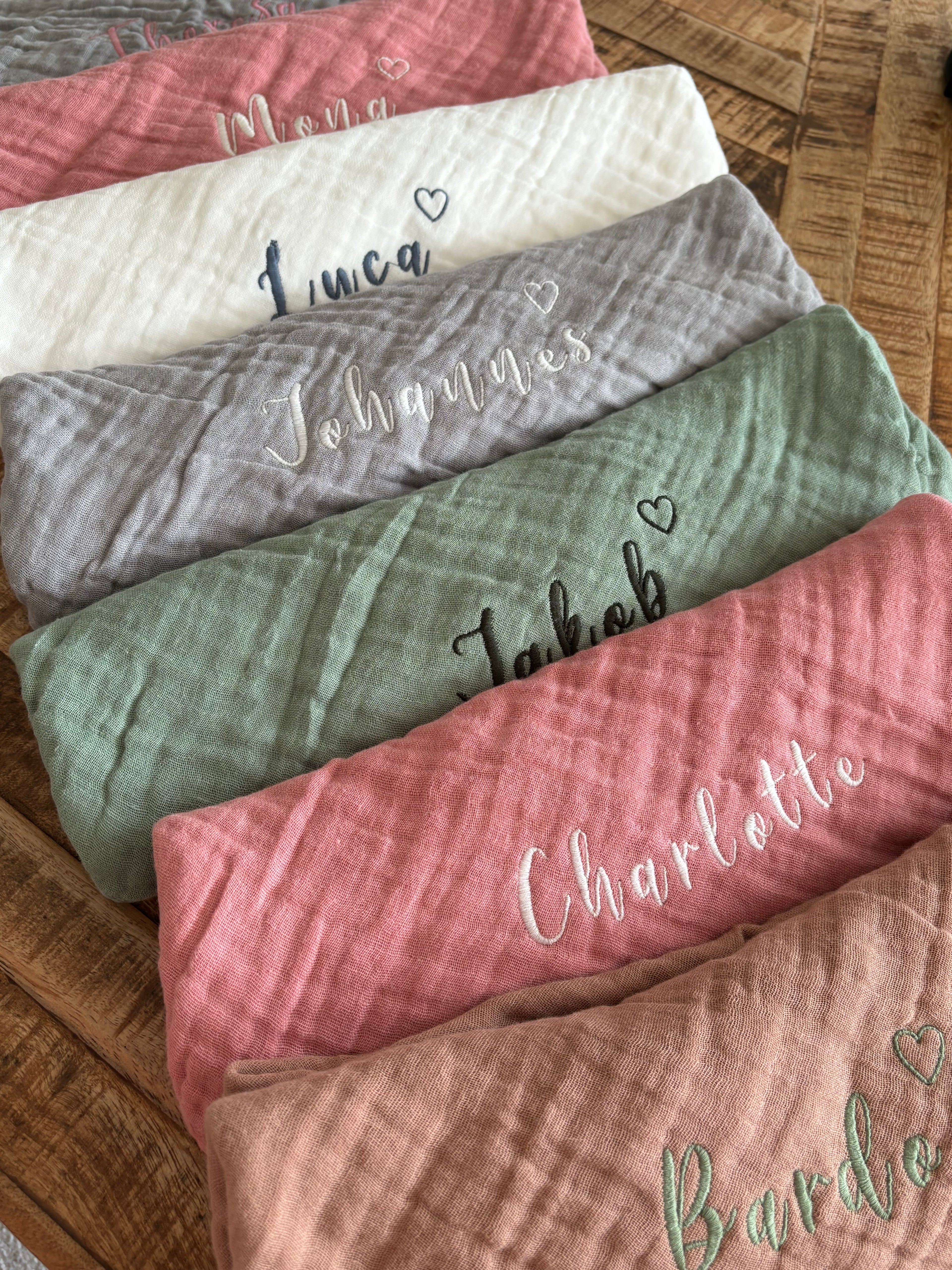 Es sind verschiedene Farben der Musselinstoff Spucktücher abgebildet. Auf jedem Tuch ist eine Namensstickerei, welche individuell und personalisiert abgestimmt ist. Zusätzlich zum Namen ist ein Herz gestickt.