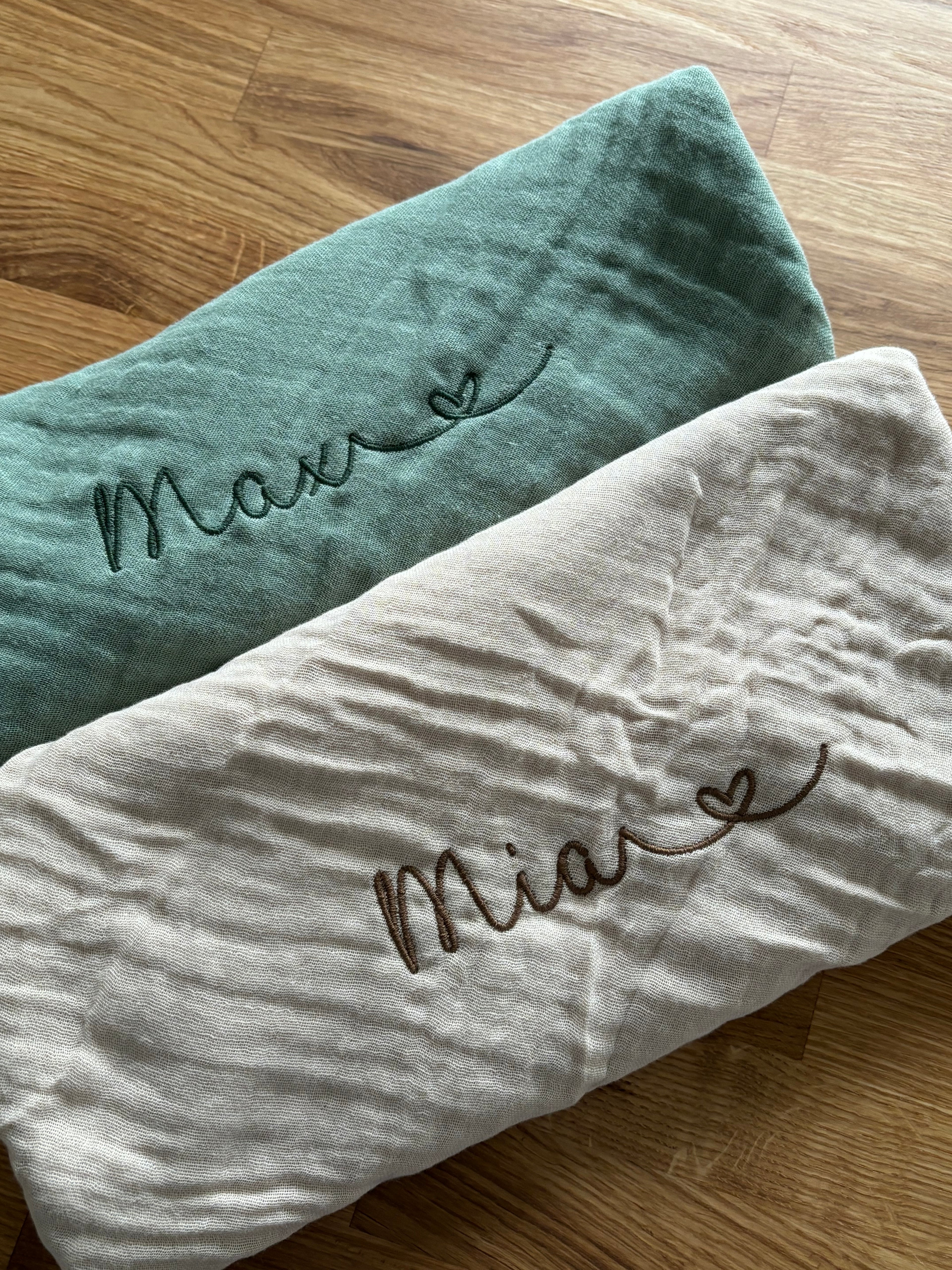 Es sind zwei Musselin Spucktücher abgebildet. Auf jedem Tuch ist eine Namensstickerei, welche individuell und personalisiert abgestimmt ist. Zusätzlich zum Namen ist ein Herz gestickt.