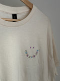 Naturales T-Shirt mit Rundhalsausschnitt und kurzen Ärmeln. Das Shirt hat ein lächelndes Gesichtsdesign, das in Regenbogenfarben gestickt ist, wobei die Worte "COOL AUNTS CLUB" das Lächeln bilden.