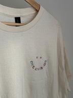 Naturales T-Shirt mit Rundhalsausschnitt und kurzen Ärmeln. Das Shirt hat ein lächelndes Gesichtsdesign, das in Regenbogenfarben gestickt ist, wobei die Worte "COOL AUNTS CLUB" das Lächeln bilden.