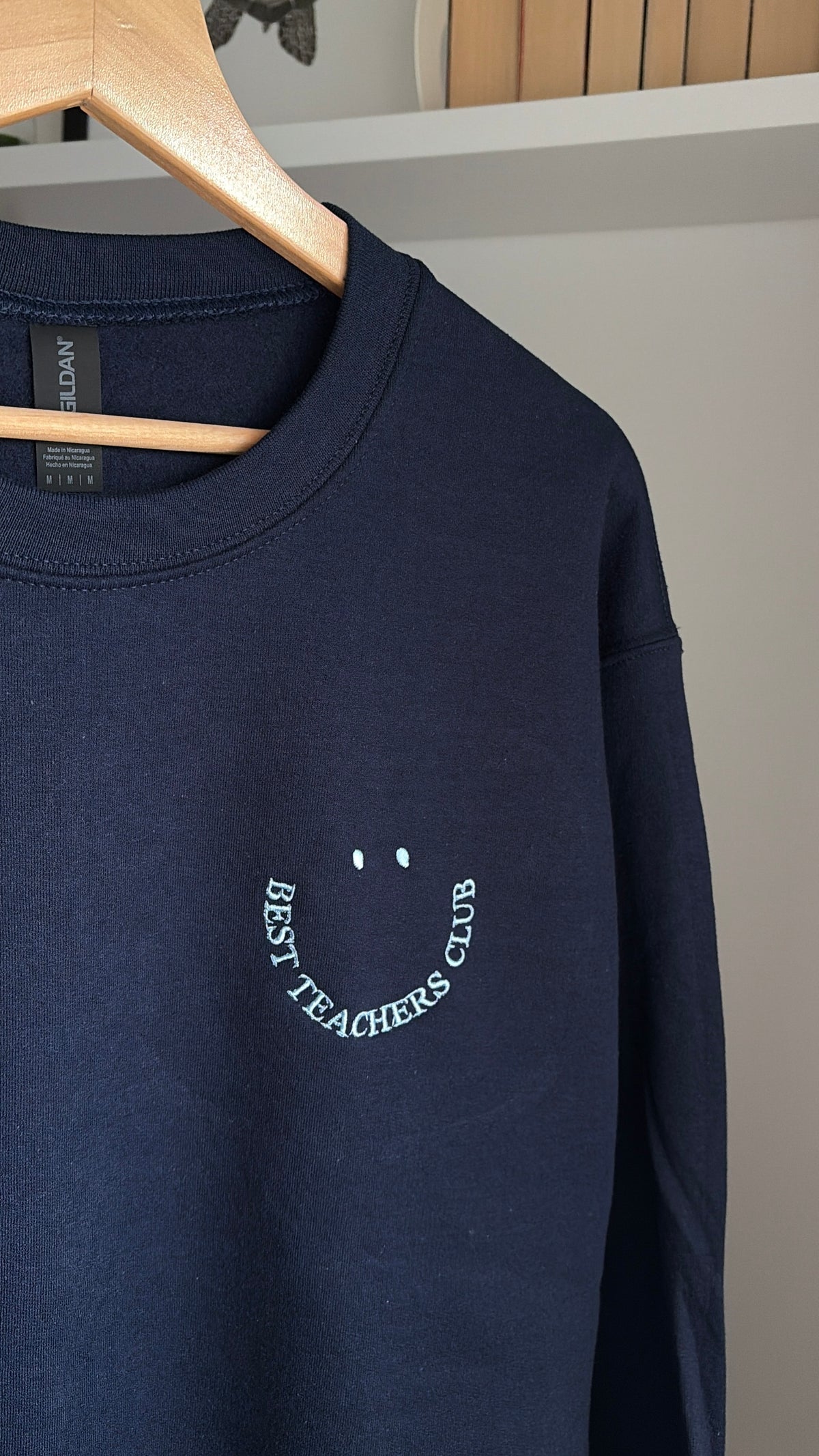 Ein marineblaues Sweatshirt mit Rundhalsausschnitt und einer weißen Stickerei auf der linken Brust. Das Motiv besteht aus zwei kleinen Punkten über einer geschwungenen Linie, die ein lächelndes Gesicht bilden, mit dem Text BEST TEACHERS CLUB.