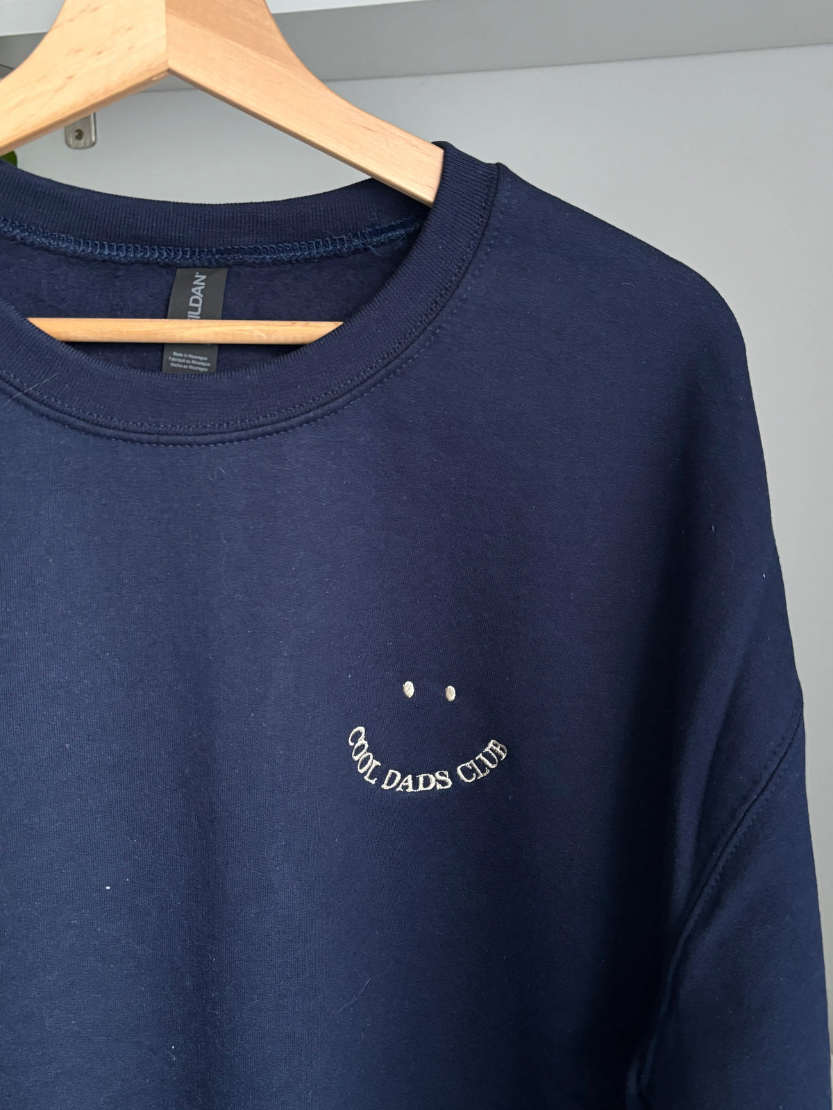 Ein Sweater in der Farbe Navy Blue mit einer beigen Stickerei vorne auf der Brust in der Form eines Smileys: Cool Dads Club.