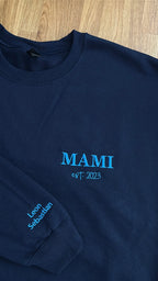 Ein Sweater in der Farbe Navy Blue mit einer Stickerei in der Farbe Aqua: Mami est. 2023. Auf dem rechten Ärmel sind zwei Kindernamen gestickt: Leon und Sebastian. 