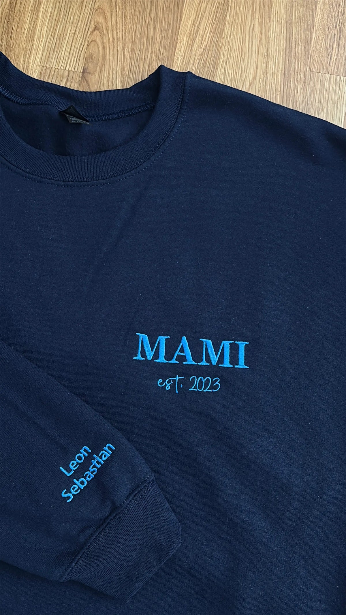 Ein Sweater in der Farbe Navy Blue mit einer Stickerei in der Farbe Aqua: Mami est. 2023. Auf dem rechten Ärmel sind zwei Kindernamen gestickt: Leon und Sebastian. 