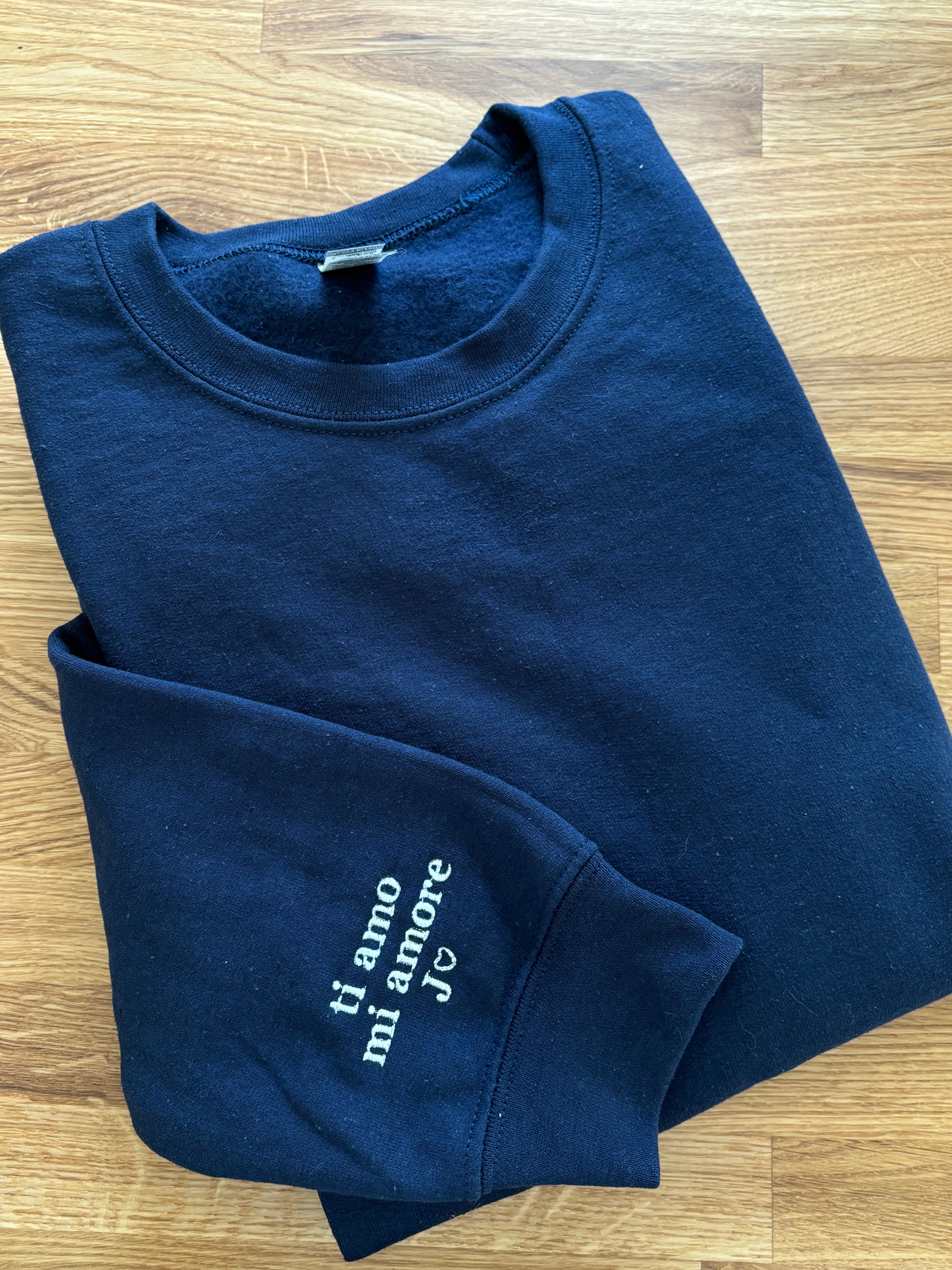 EIn Sweater in der Farbe Navy Blue auf einem Holztisch. Auf dem Ärmel ist in weiß "ti amo mi amore J" gestickt.
