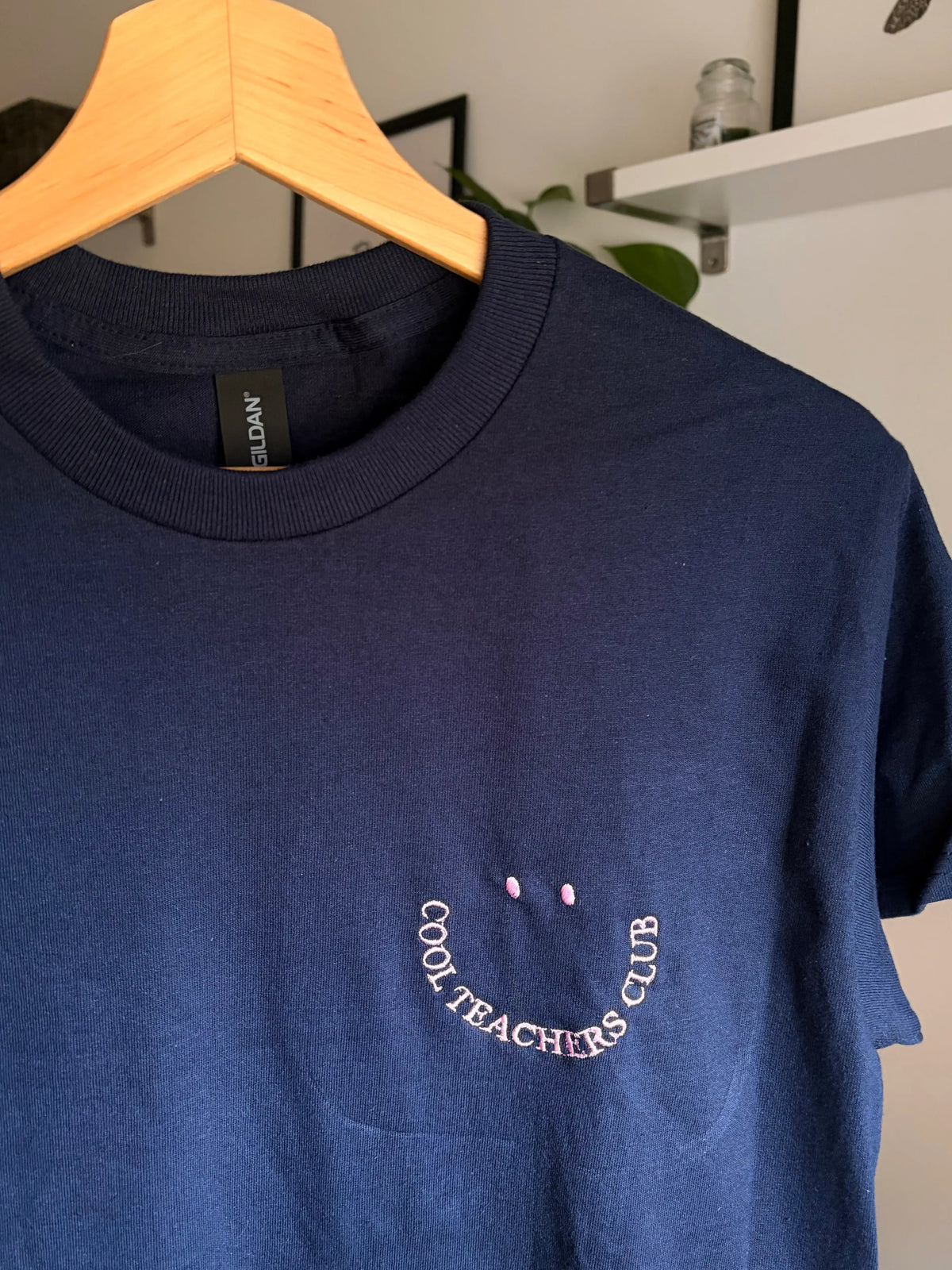 Ein T-Shirt in der Farbe Navy Blue mit einem rosa  Stickerei vorne links auf der Brust „Cool Teachers Club" in Großbuchstaben.