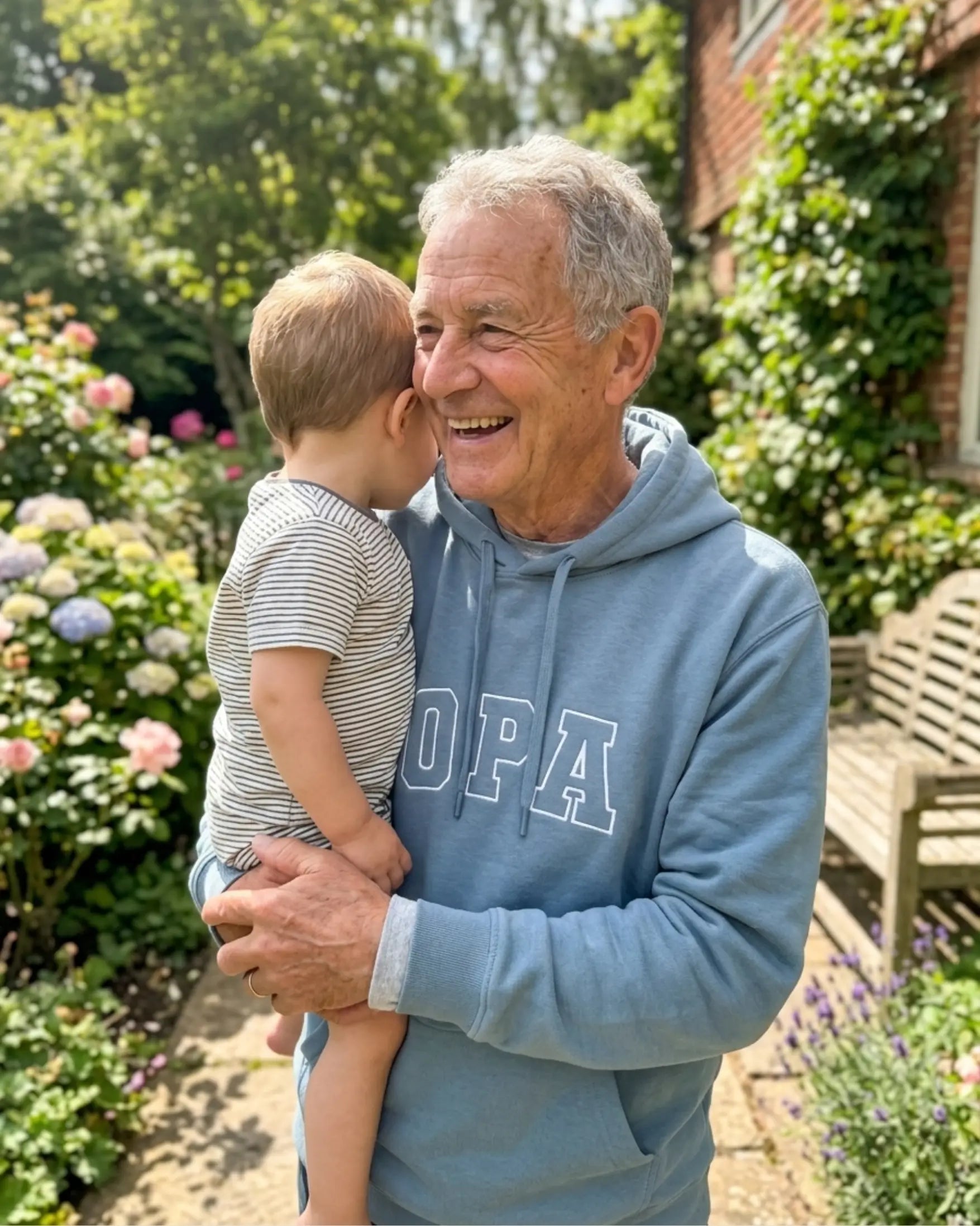 Ein lächelnder älterer Mann in einem hellblauen Kapuzenpullover mit der Aufschrift „OPA“ steht in einem sonnigen Garten und hält ein Kleinkind auf dem Arm. Das Kind trägt ein gestreiftes T-Shirt und blickt vom Betrachter weg über die Schulter des Mannes. Im Hintergrund sind blühende Blumen und ein Backsteinhaus zu sehen.