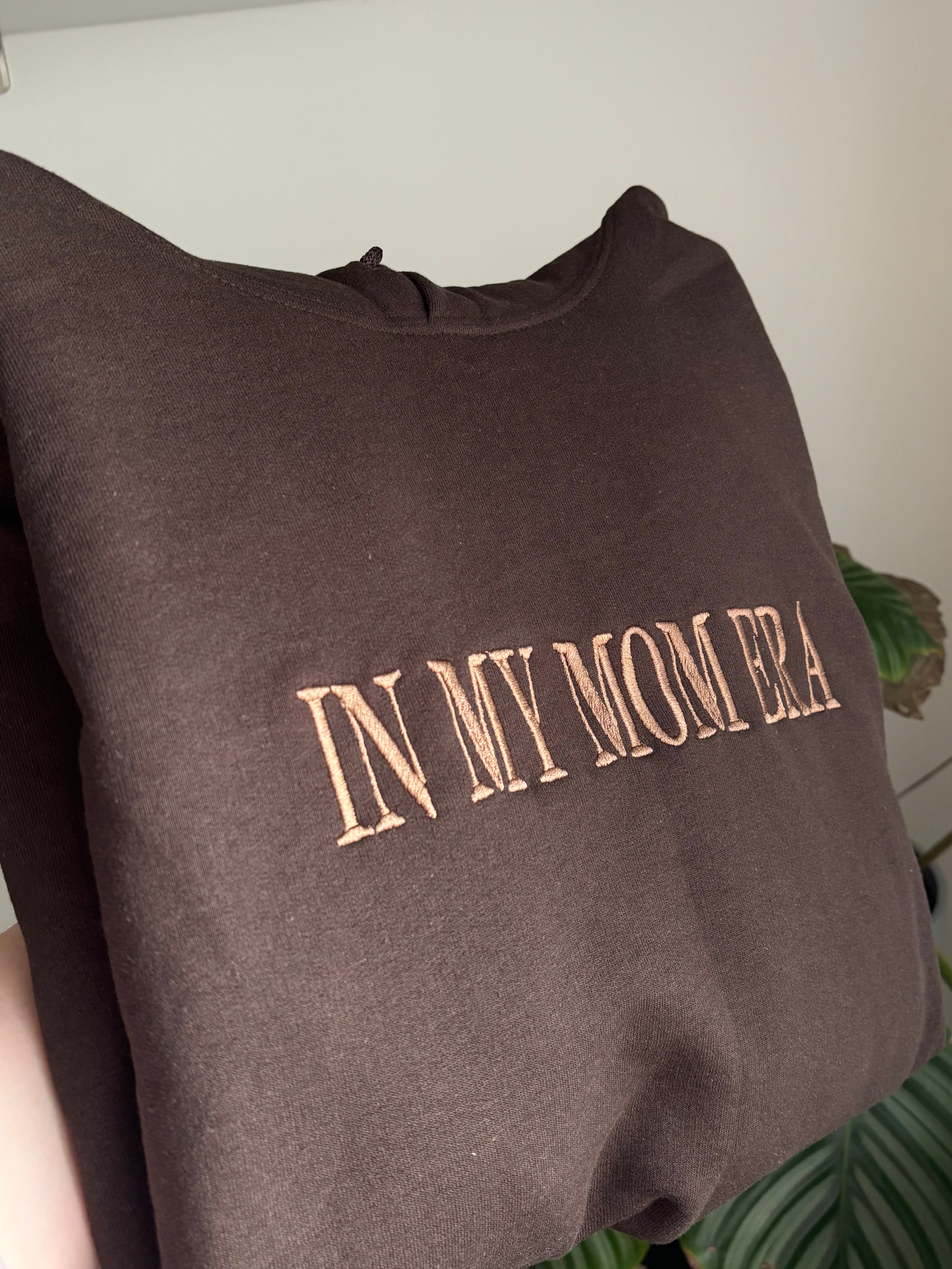 Ein Hoodie in der Farbe Dark Chocolate mit dem Wunschtext „IN MY MOM ERA“ in der Garnfarbe Brown.