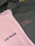 Zwei Hoodies in rosa und dunkelgrau mit einer Stickerei jeweils auf der linken Seite: I'm Preace und I come in peace.