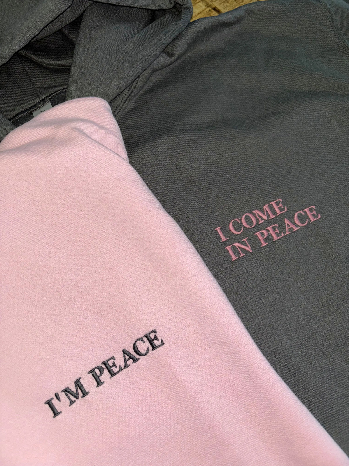 Zwei Hoodies in rosa und dunkelgrau mit einer Stickerei jeweils auf der linken Seite: I'm Preace und I come in peace.