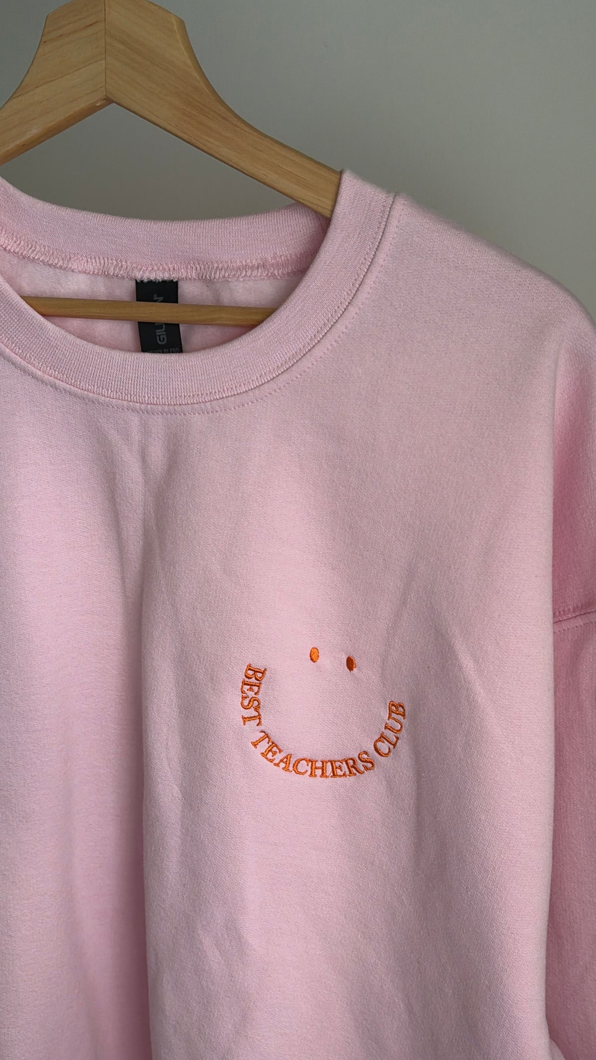 Ein hellrosa Sweatshirt mit Rundhalsausschnitt und orangefarbener Stickerei auf der Brust. Die Stickerei zeigt ein Smiley-Gesicht, das aus zwei kreisförmigen Augen und einer geschwungenen Textzeile "BEST TEACHERS CLUB" in serifenloser Schrift besteht.