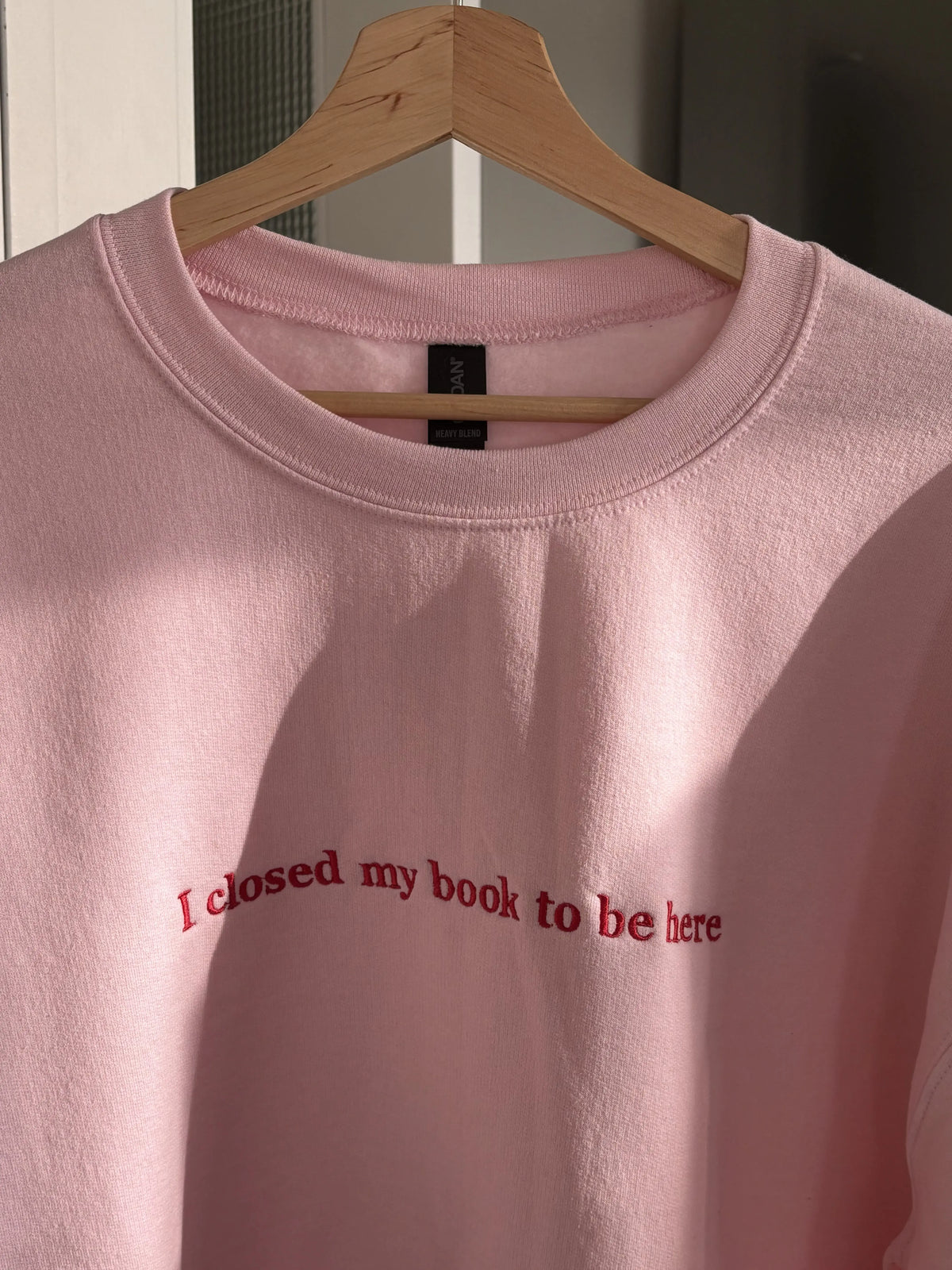 Ein rosa Sweater mit dem Schriftzug I closed my Book To be Here. Der Sweater ist mit einem roten Garn bestickt.