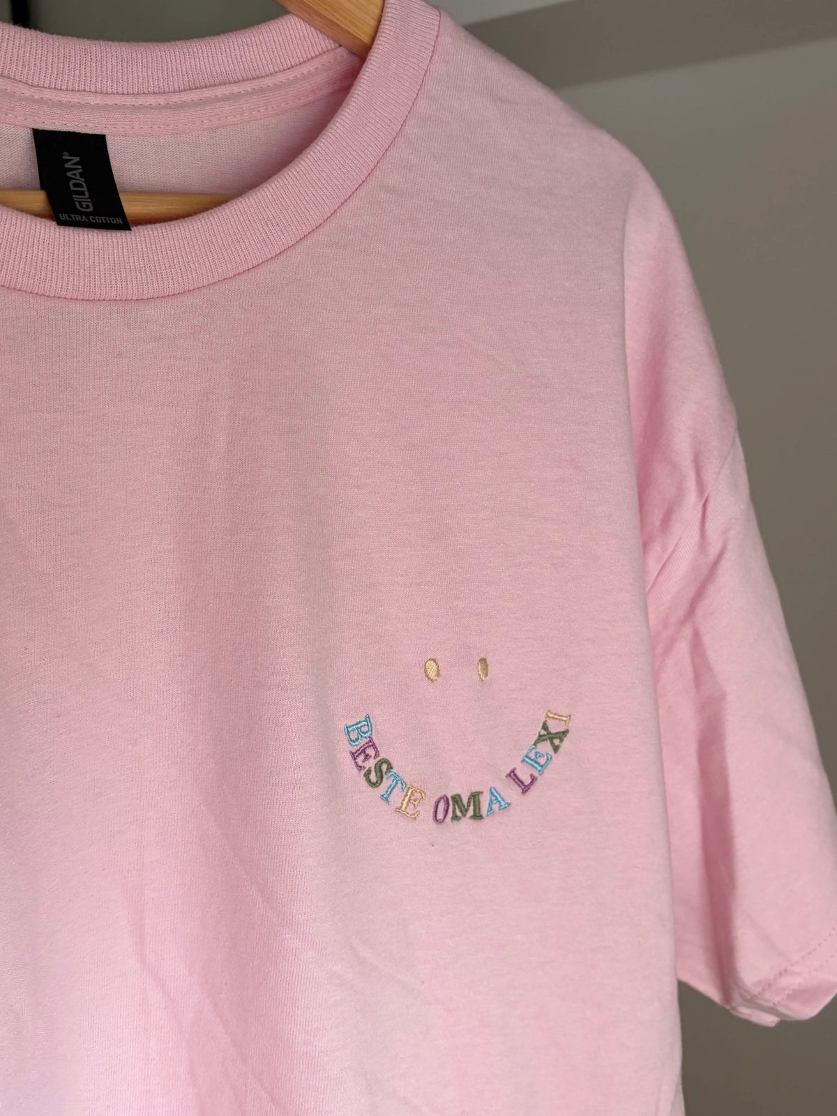 Ein T-Shirt in der Farbe Rosa mit kurzen Ärmeln und einen bunten Smiley auf der linken Brustseite. Die Grafik besteht aus zwei kleinen ovalen Augen und einem geschwungenen Schriftzug BESTE OMA LEXI, der ein lächelndes Gesicht bildet.