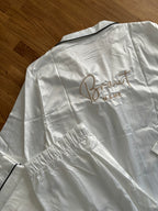 Ein weißer Pyjama aus Satin. Auf dem Rücken ist in Beige Braut Name gestickt. Der Pyjama liegt auf einem Holztisch. 