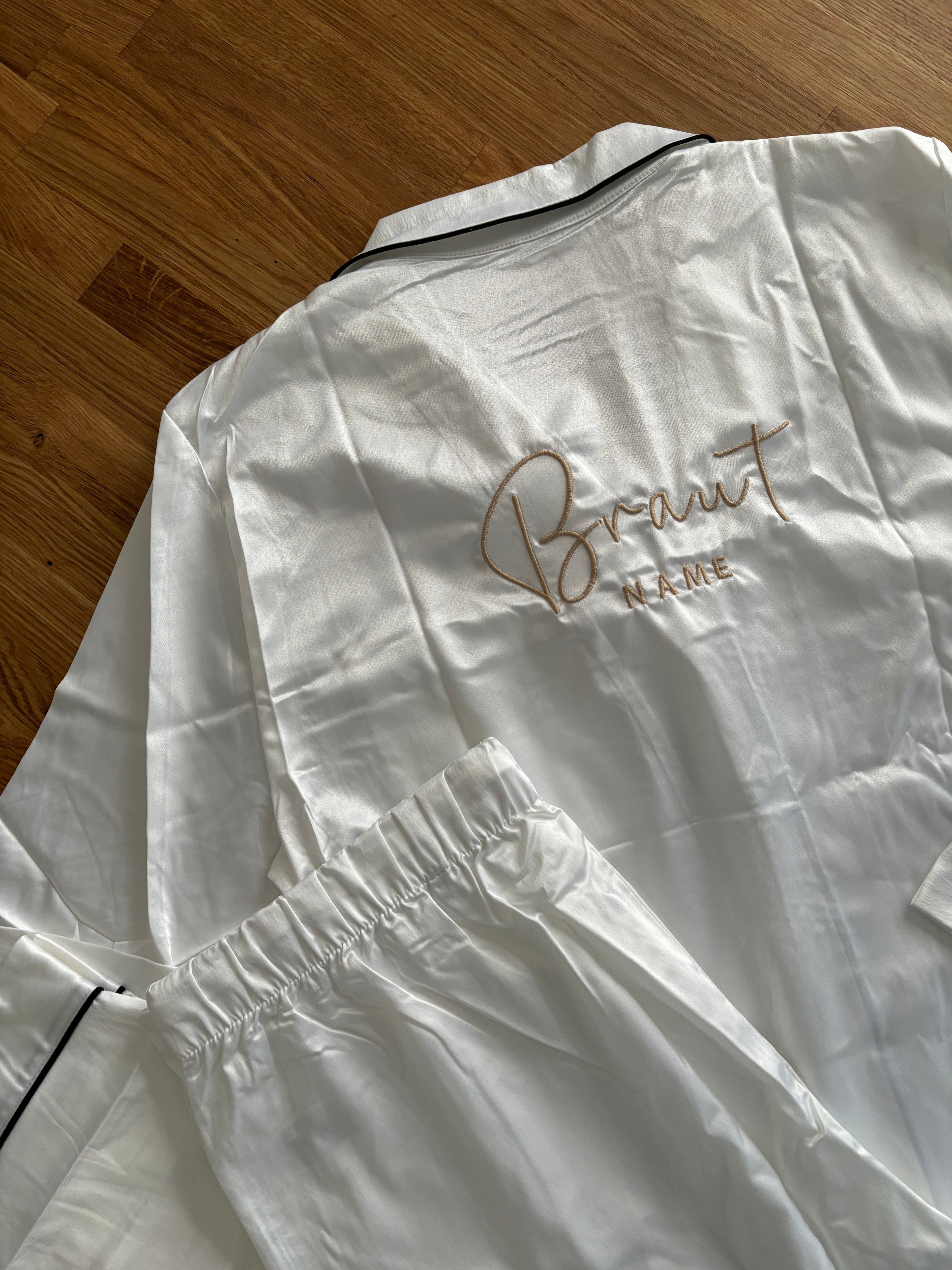 Ein weißer Pyjama aus Satin. Auf dem Rücken ist in Beige Braut Name gestickt. Der Pyjama liegt auf einem Holztisch. 