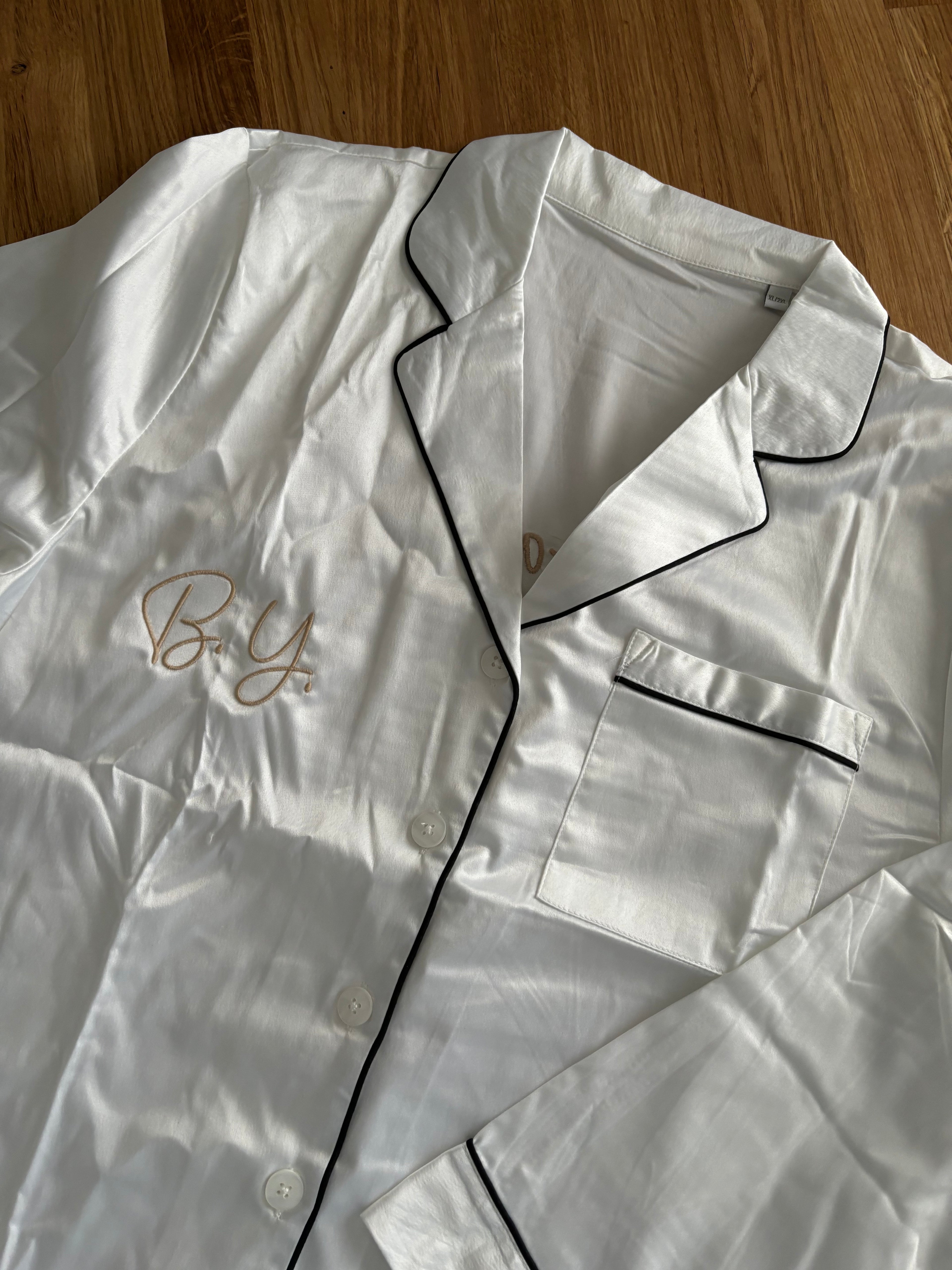 Das Oberteil vom Pyjama ist weiß und liegt auf einem Holztisch. Der Pyjama hat eine schwarze Umrandung. Vorne ist B.Y. gestickt.