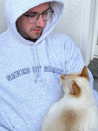 Ein Mann trägt einen Kapuzen Hoodie mit dem Stick vorne "Saved By Grace" in Großbuchstaben und in Blau. Der Mann trägt eine Katze auf dem Arm.