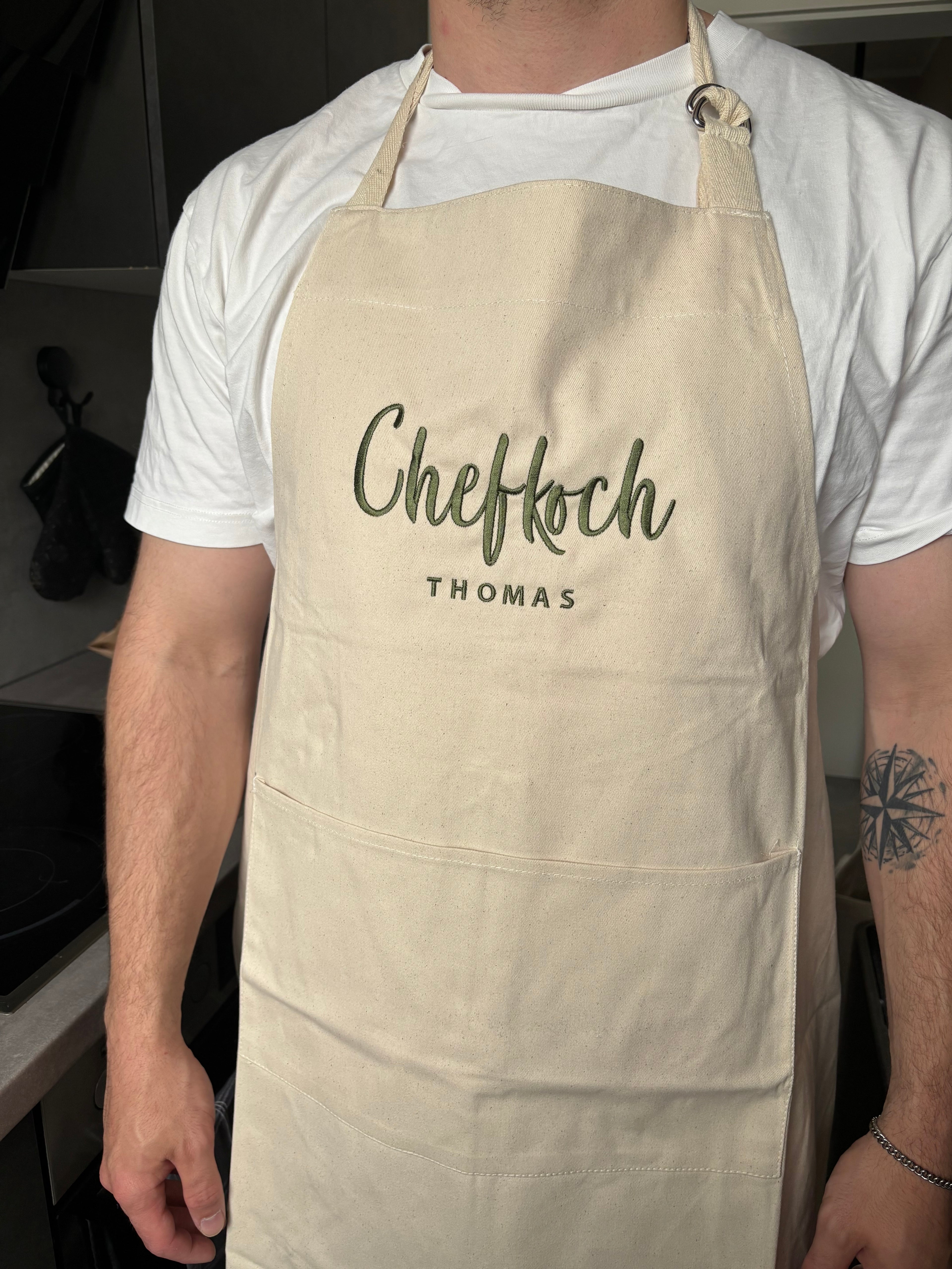 Ein Mann, der eine personalisierte Schürze trägt. Auf der Kochschürze ist in grün "Chefkoch Thomas" gestickt.