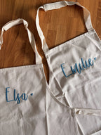 Zwei Kochschürzen für Kinder für ein Geschwisterpaar. Vorne mittig ist jeweils der Kindername in blau mit einem Herz gestickt: Elsa und Emilie.