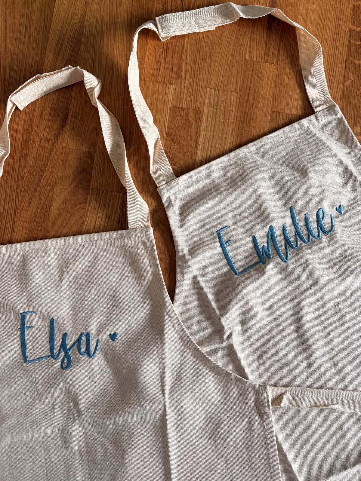 Zwei Kochschürzen für Kinder für ein Geschwisterpaar. Vorne mittig ist jeweils der Kindername in blau mit einem Herz gestickt: Elsa und Emilie.