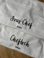 Hier sind zwei Kochschürzen zu sehen, auf der einen steht Chefkoch Julian und auf der anderen Sous Chef Anton. Die Garnfarbe ist coffee Brown und hat eine geschwungene Schrift.