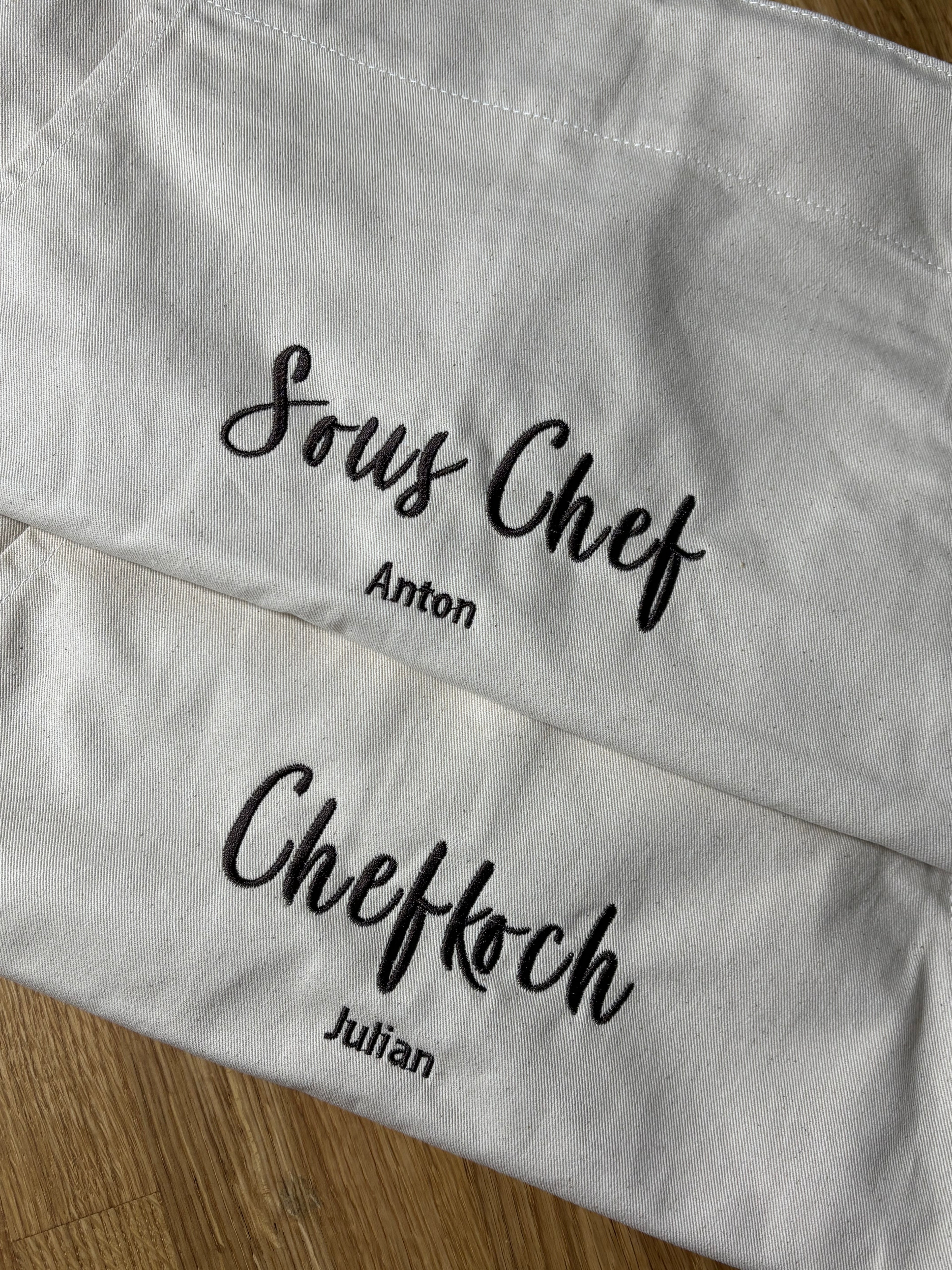 Hier sind zwei Kochschürzen zu sehen, auf der einen steht Chefkoch Julian und auf der anderen Sous Chef Anton. Die Garnfarbe ist coffee Brown und hat eine geschwungene Schrift.