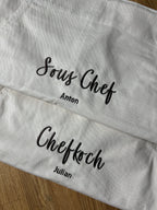 Hier sind zwei Kochschürzen zu sehen, auf der einen steht Chefkoch Julian und auf der anderen Sous Chef Anton. Die Garnfarbe ist coffee Brown und hat eine geschwungene Schrift.