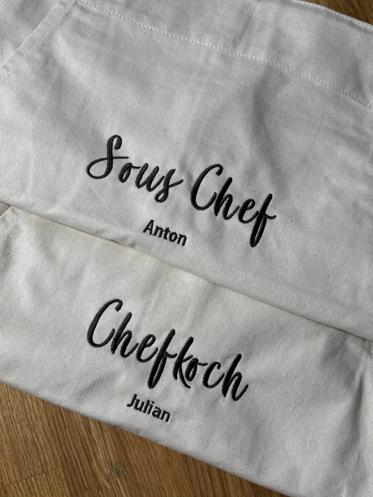 Hier sind zwei Kochschürzen zu sehen, auf der einen steht Chefkoch Julian und auf der anderen Sous Chef Anton. Die Garnfarbe ist coffee Brown und hat eine geschwungene Schrift.