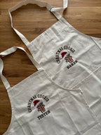 Zwei Schürzen für Kinder und Erwachsene liegen auf einem Holztisch. Auf den Schürzen ist jeweils mittig ein Cookie gestickt: Christmas Cookie Tester und Baker.