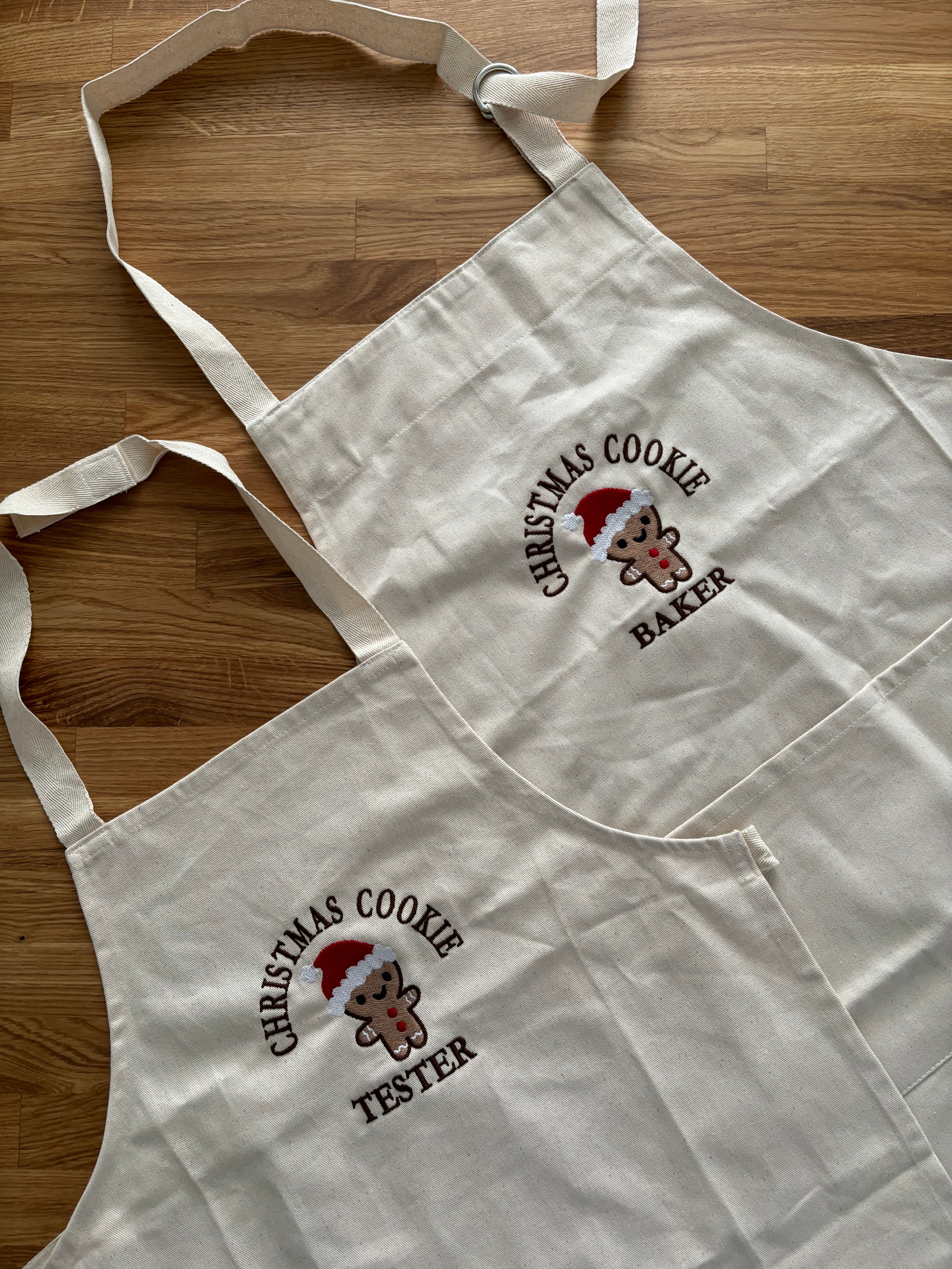 Zwei Schürzen für Kinder und Erwachsene liegen auf einem Holztisch. Auf den Schürzen ist jeweils mittig ein Cookie gestickt: Christmas Cookie Tester und Baker.
