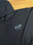 Ein Hoodie in der Farbe schwarz mit einer Stickerei in der Farbe Light Blue: Dad est. 2026.