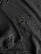 Ein Hoodie in der Farbe Schwarz mit einer Stickerei in der Farbe Schwarz: Dad. Auf dem rechten Ärmel ist ein Kindername gestickt: Jakob. 