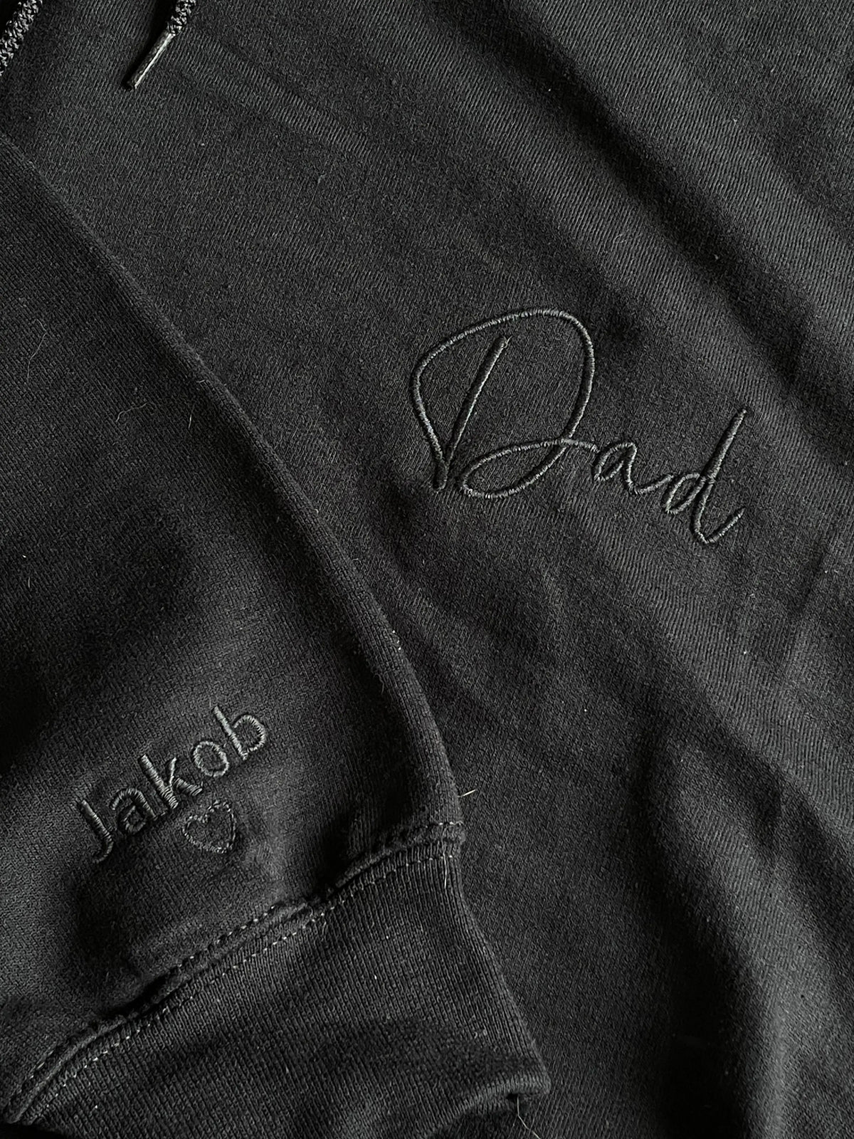 Ein Hoodie in der Farbe Schwarz mit einer Stickerei in der Farbe Schwarz: Dad. Auf dem rechten Ärmel ist ein Kindername gestickt: Jakob. 