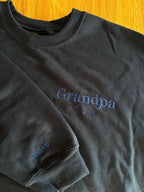 Ein Sweater in der Farbe Schwarz mit einer Stickerei in der Farbe Navy Blue: Grandpa est. 2024. Auf dem rechten Ärmel ist der Kindername gestickt: Julia. 