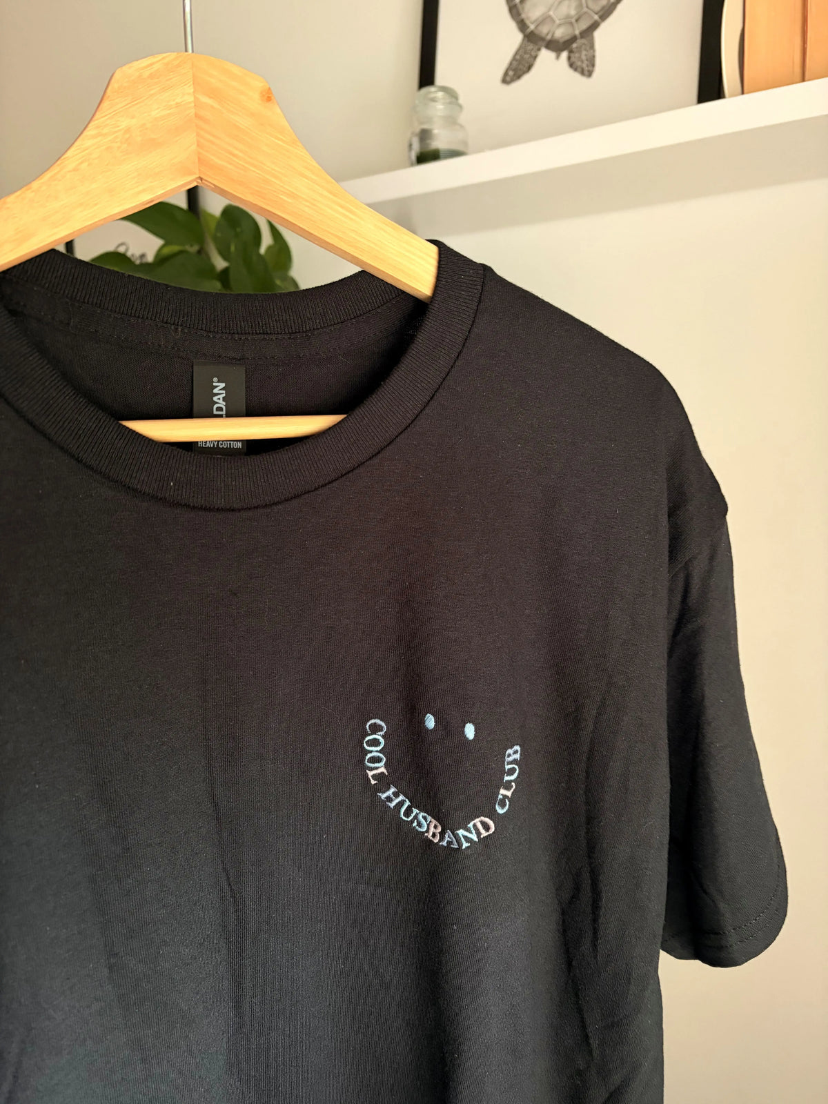 Ein T-Shirt in der Farbe Schwarz mit einem Smiley bestickt "COOL HUSBAND CLUB".
