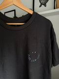 Ein schwarzes T-Shirt mit Rundhalsausschnitt und einer Stickerei auf der linken Brust. Das Design besteht aus zwei kleinen grauen Punkten als Augen über einer geschwungenen Linie aus mehrfarbigem Text, der COOL UNCLES CLUB bildet.
