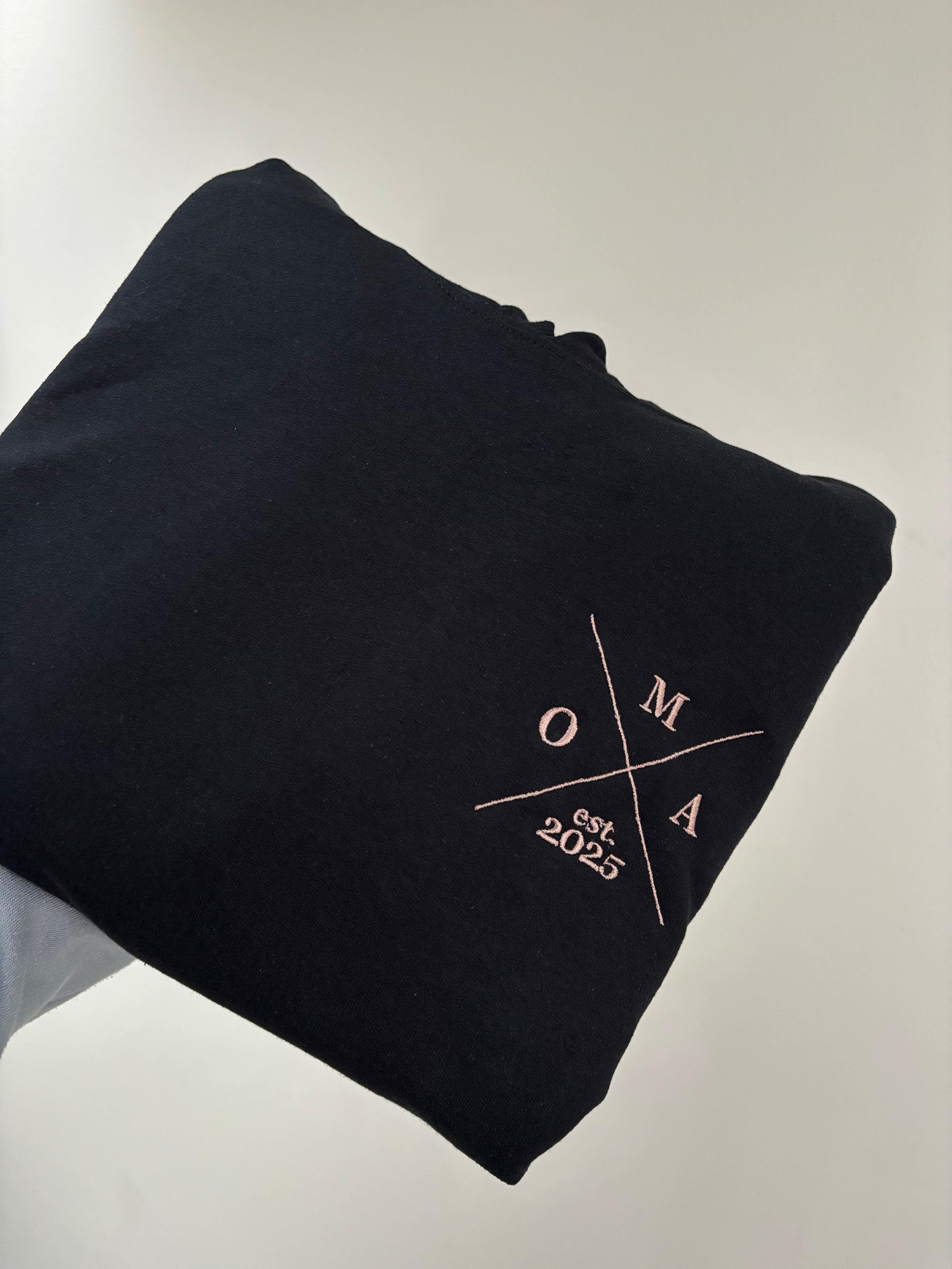 Ein Hoodie in der Farbe Schwarz mit einem Koordinaten Kreuz auf der linken Brust gestickt. Im Kreuz steht Oma est. 2025.
