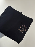 Ein Hoodie in der Farbe Schwarz mit einem Koordinaten Kreuz auf der linken Brust gestickt. Im Kreuz steht Oma est. 2025.