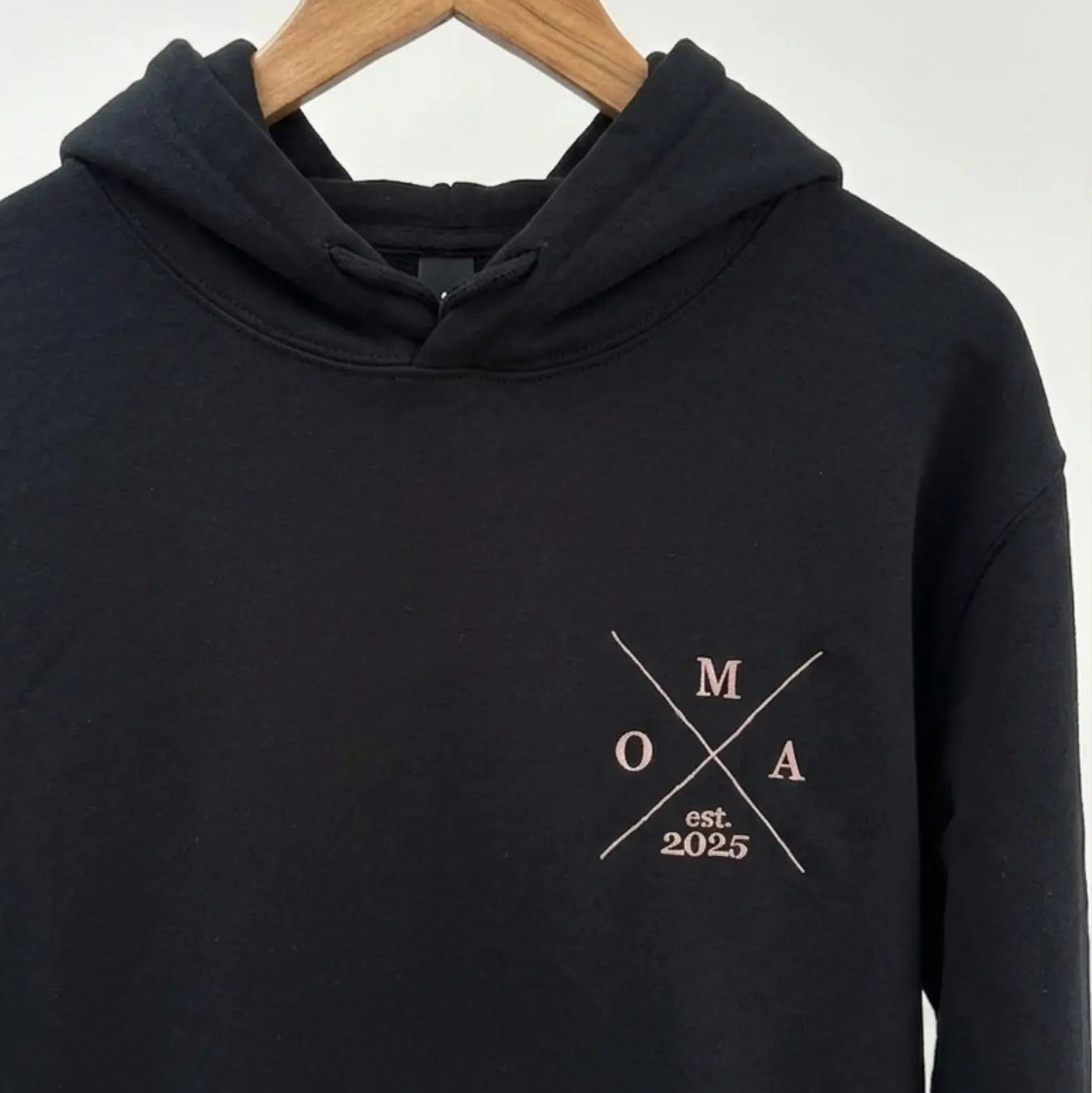 Ein schwarzer Hoodie mit Kapuze auf einem Holzbügel. Der Pullover ist vorne bestickt: Oma est. 2025.