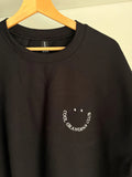 Schwarzer Sweater und einer weißen Smiley-Gesicht-Stickerei mit der Aufschrift "COOL GRANDPAS CLUB". Der Pullover hängt an einem Holzkleiderbügel.