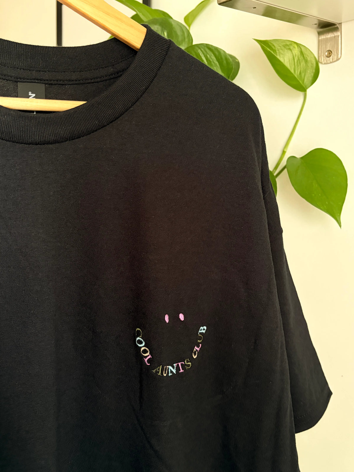 Schwarzes T-Shirt mit Rundhalsausschnitt. Das Shirt hat ein gesticktes Smiley-Gesicht und die Worte "COOL AUNTS CLUB" in einem geschwungenen Design. Der Text und das Smiley-Gesicht sind in verschiedenen Farben.