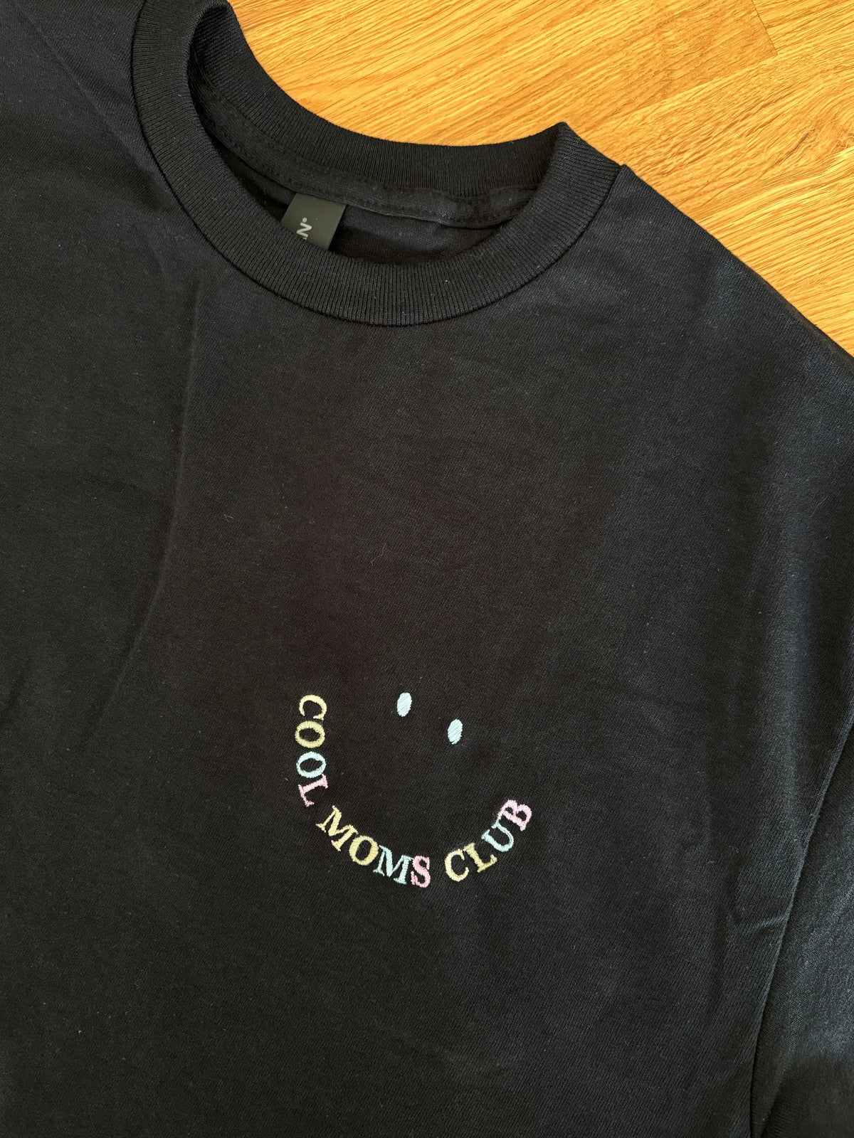Ein schwarzes T-Shirt mit einem Smiley bestickt "COOL MOMS CLUB".