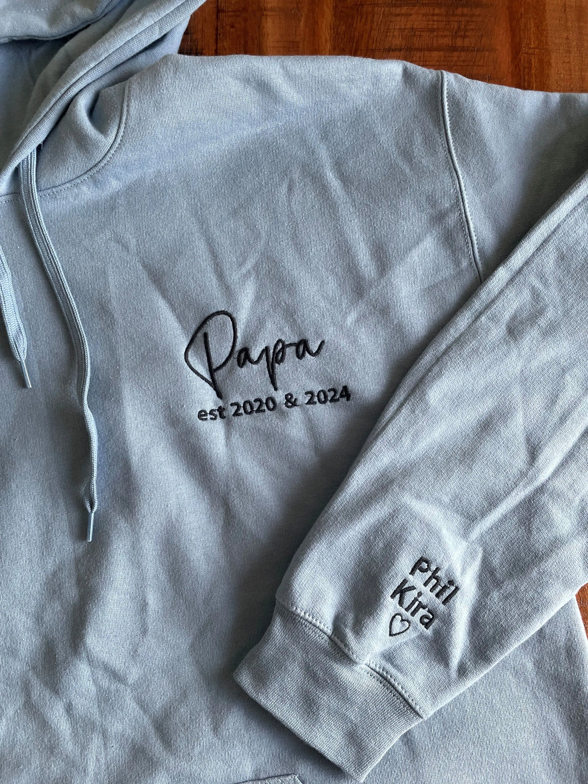 Ein Hoodie in der Farbe Stone Blue mit einer Stickerei in der Farbe Schwarz: Papa est. 2020 & 2024. Auf dem linken Ärmel sind zwei Kindernamen gestickt: Phil und Kira. 