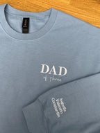 Ein Sweater in der Farbe Stone Blue. Der Stick ist in weiß. Gestickt ist in Großbuchstaben DAD und darunter of three. Auf dem Ärmel stehen drei Kindernamen: Isabella, Valentin und Constantin.