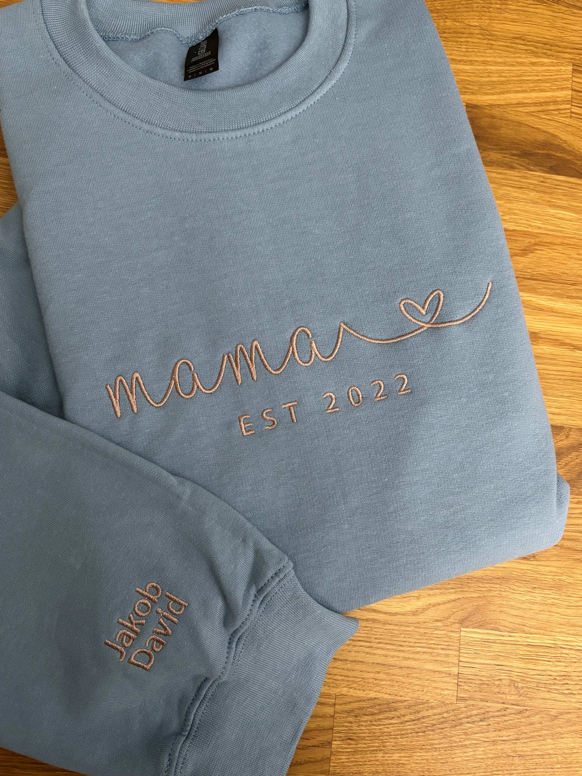 Ein Hoodie in der Farbe Stone Blue mit einer Stickerei in beige auf der Brust: Mama. Zusätzlich sind die Kindernamen auf dem Ärmel gestickt.