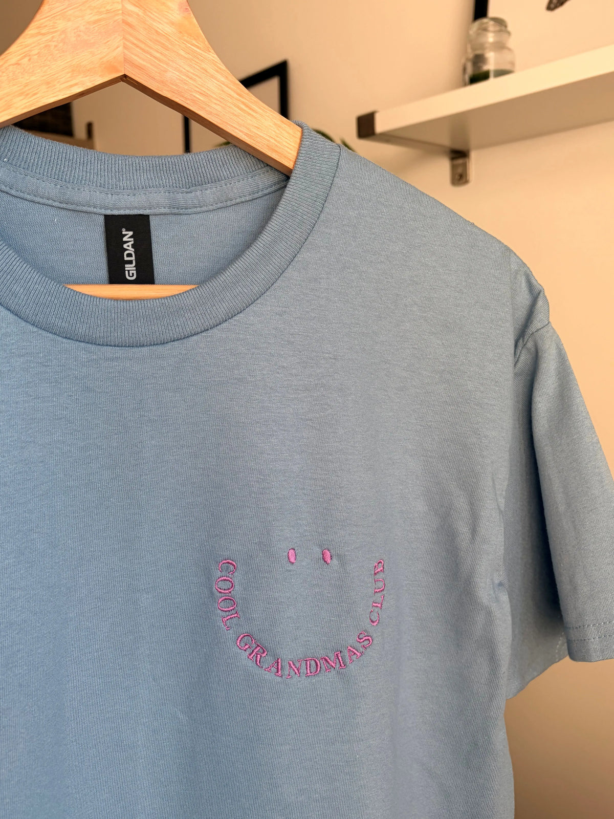 Ein T-Shirt in der Farbe Stone Blue mit kurzen Ärmeln und einen lila Smiley auf der linken Brustseite. Die Grafik besteht aus zwei kleinen ovalen Augen und einem geschwungenen Schriftzug COOL GRANDMAS CLUB, der ein lächelndes Gesicht bildet.