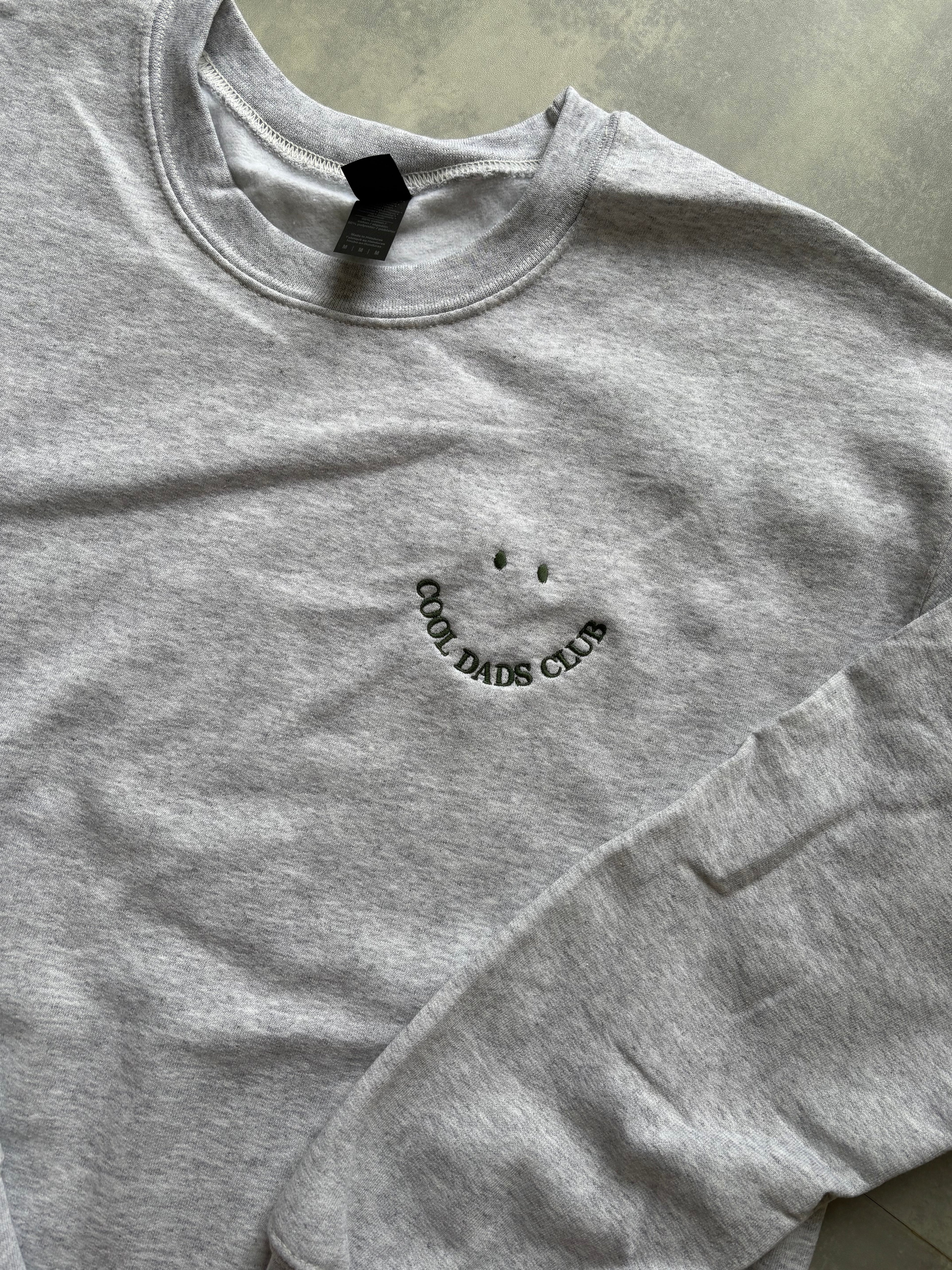Ein Sweater in der Farbe Ash Heather mit einer dunkel grünen Stickerei Cool Dads Club.
