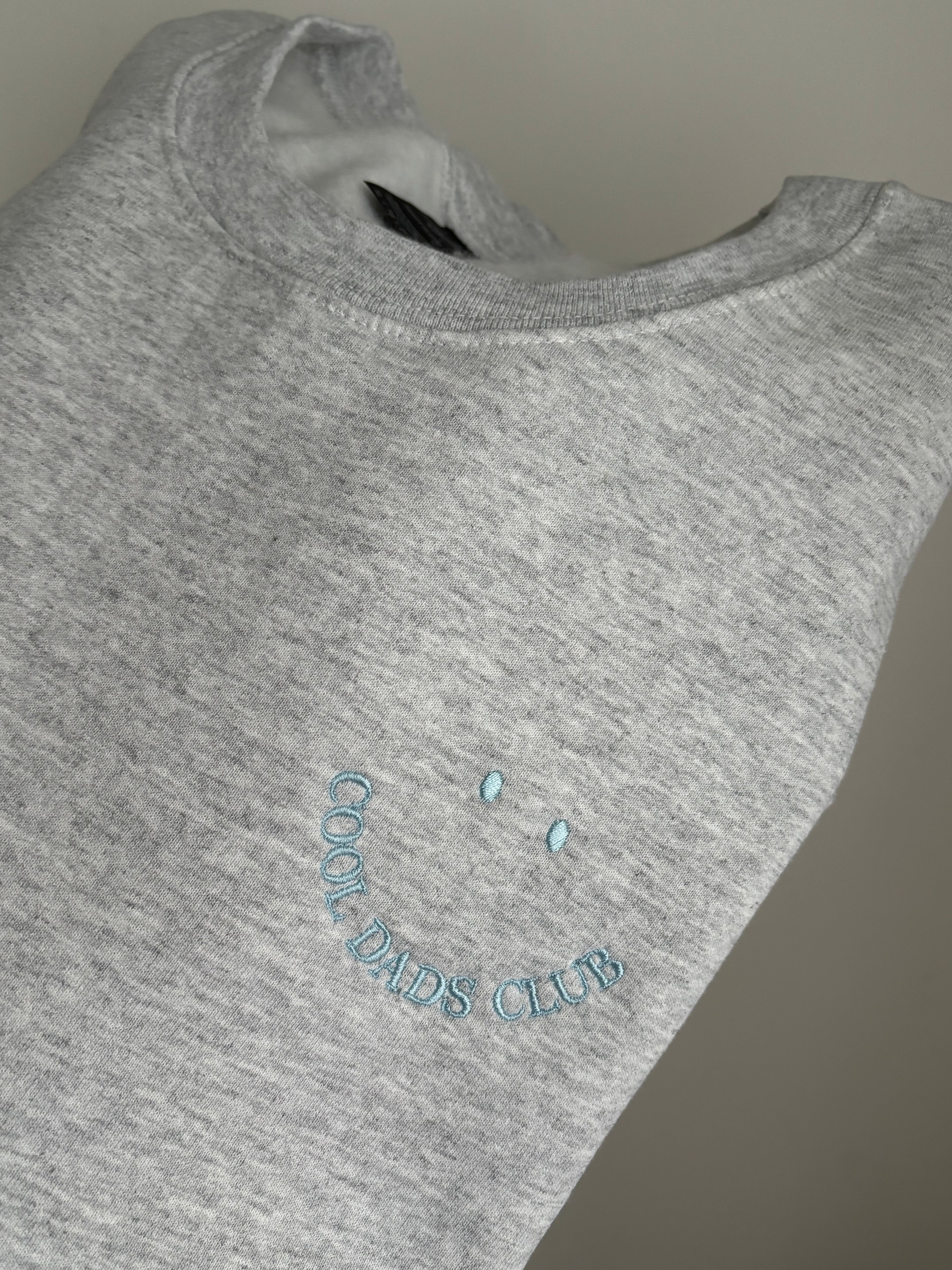 Ein Sweater in der Farbe Ash Heather mit einer hellblauen Stickerei Cool Dads Club.