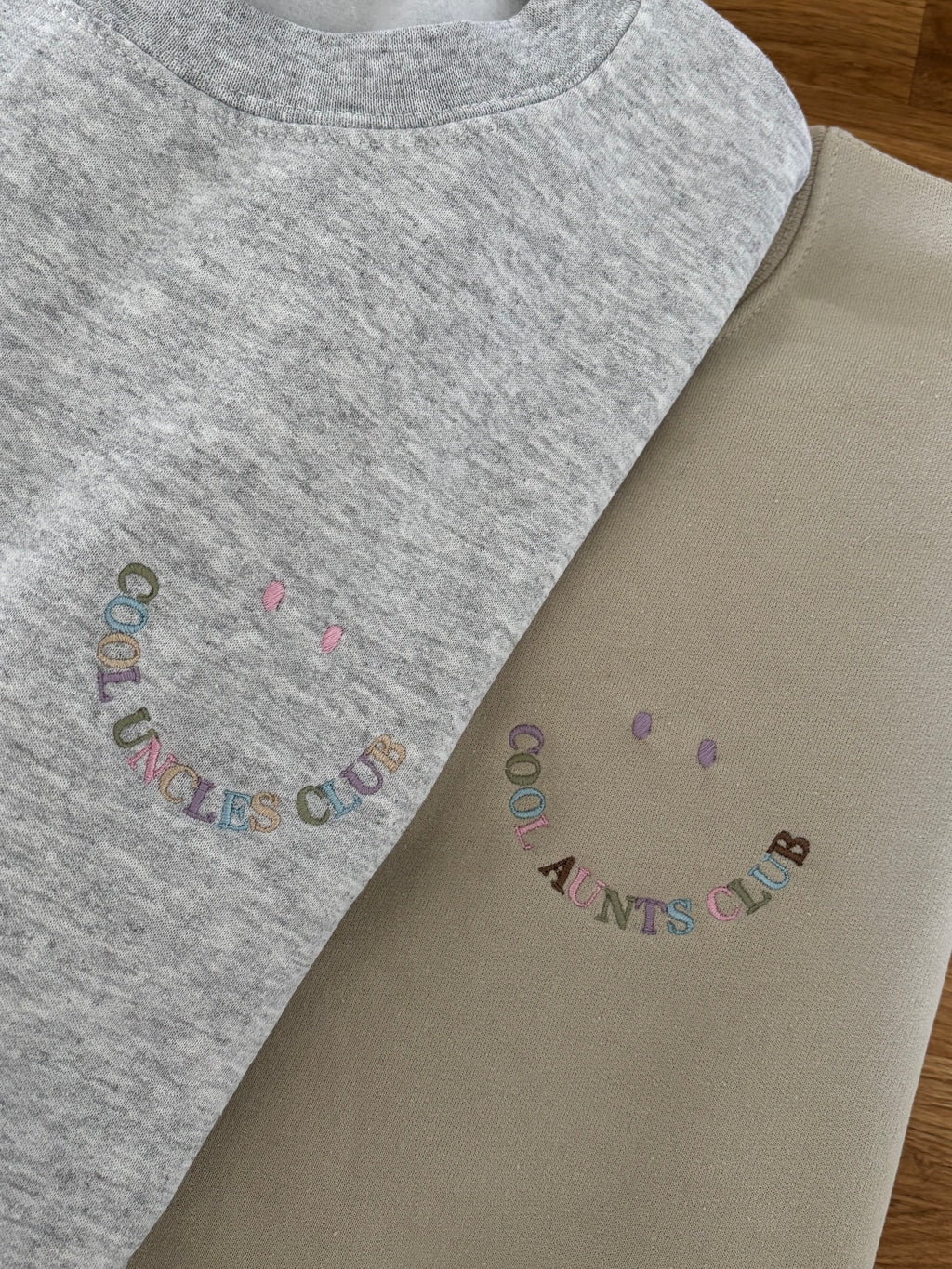 Zwei Sweater in beige und ash heather mit jeweils einer Stickerei, die wie ein Smiley aussieht: Cool Uncles Club und Cool Aunts Club.